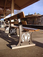 A bench seen from low down and behind. Three brown-painted cast iron legs have 