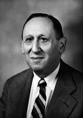 Head and shoulders of a man in his early 60s in coat and tie, facing slightly to his right. He is balding and has a serious but slightly smiling expression.