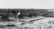 Foreground, a battery of 16 heavy guns. Background, conical tents and support vehicles.