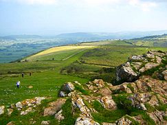 Rocks in front of grassy open spaces with walkers. Hills in the distance