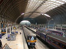 The interior of a large railway station with a curved roof supported by iron girders, supported by iron columns, four diesel trains standing at platforms, passengers on the platforms, in the distance daylight can be seen and the scene is illuminated by natural light through the centre section of the roof