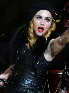 A middle-aged blond woman smiling towards the camera. She wears bright, red lipstick and a black dress with lots of straps and fur lining in it. The woman wears a cap, from which her hair comes down in waves. Her hands are near her face and she holds a microphone in her right one.