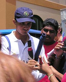 A middle-aged man signing on cricket bats. He wears a white t-shirt and a navy blue cap. A number of people are visible, who surround him.