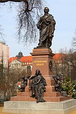 coloured photograph of a statue of a robed male figure on a  stepped pedestal inscribed 'Felix Mendelssohn Bartholdy', with a seated female figure holding a lyre at its base, situated in an open space