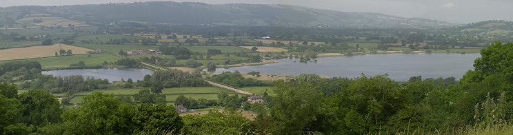 photograph of the lake showing a road crossing via a causeway at Herriots Bridge. It is surrounded by green vegetation with hills in the distance