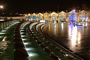 Opinión de la noche a través de una plaza abierta dominada por una fuente de agua larga curva que es decorativamente lit. En el otro extremo de la plaza se encuentra la fachada de un edificio abovedado estación de tren