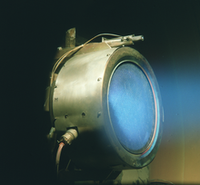 Un cilindro de metal con electrodos conectados a su lado. Luz difusa Azul está saliendo del tubo.