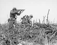 una imagen en blanco y negro de dos infantes de marina en sus uniformes de combate. Un marine es proporcionar fuego de cobertura con su metralleta Thompson M1 como el otro con un fusil automático Browning, se prepara para romper la cubierta para moverse a una posición diferente. Hay palos desnudos y rocas en el suelo.