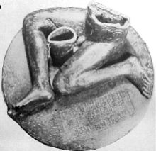 Fotografía en blanco y negro de una estatua que consiste en una inscripción, pedestal redondo encima de los cuales se sienta una figura desnuda, masculina sedente de la que se conservan sólo las piernas y la parte inferior del torso
