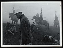 Kurosawa, un hombre alto y delgado, a principios de la edad media frente a la izquierda, vistiendo ropa moderna coronadas por una gorra blanca y suave, paseos a través de un paisaje de niebla, como detrás de él varios actores en atuendo medieval japonesa parecen a caballo o acostado o sentado en el suelo.