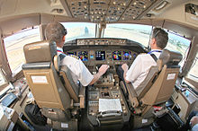 Una cubierta de vuelo, desde detrás de los asientos de los dos pilotos. Una consola central se encuentra entre los asientos, en frente es un panel de instrumentos con varias pantallas, y la luz entra a través de las ventanas delanteras.