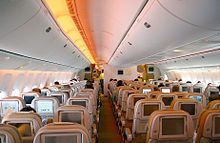 Cabina del avión de pasajeros. Filas de asientos dispuestos entre dos pasillos. Cada respaldo tiene un monitor; luz brilla desde las paredes laterales y compartimentos superiores.