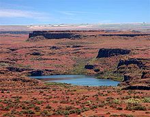 Terreno de color marrón rojizo y muchos pequeños arbustos verdes rodean un lago. Crestas truncadas de roca oscura corren a través del terreno paralelo al horizonte y entre sí bajo un cielo azul.