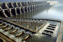 Water flows down a stair-step aqueduct from the top of a large river dam to the bottom. The aqueduct makes two 90-degree turns to the left on the way down before reaching the river at the base of the dam.