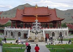 Fotografía de un gran edificio de Mongolia de techos rojos, con un árbol metálico elaborado delante de él, a unos 30 metros de altura