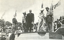 Cinco personas en la plataforma de piedra, de izquierda a derecha: El hombre con el bigote en uniforme de gala militar y el sombrero negro alto tupida con barbijo; hombre en uniforme militar con muchas medallas y el sombrero de oficial de la marina antigua usanza; hombre con el pelo blanco en juego con el abrigo largo y oscuro de cuello blanco; mujer en vestido victoriano con sombrero blanco; hombre con la barba y el pelo revuelto en traje. Multitud y banderas en el fondo.