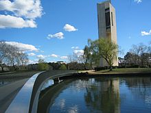 Tall edificio de color gris claro, con paredes de forma irregular y ventanas altas y estrechas se extiende hacia el cielo antes de un árbol y el lago, un puente cruza el lago para el edificio.