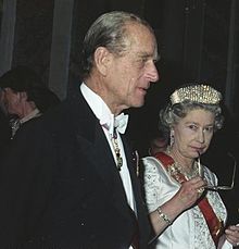 Elizabeth, en traje formal, tiene un par de gafas a la boca en una actitud pensativa