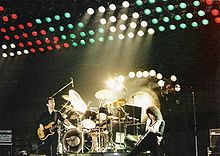 Tres miembros del grupo durante una actuación en directo en Hannover. De izquierda a derecha, John Deacon (de pie casualmente), Roger Taylor (de juego, se sentó en el kit de batería) y Brian May (parece estar jugando intensamente). Detrás de Taylor es un tam-tam utiliza al final de Bohemian Rhapsody. Detrás de eso es un gran juego de luces multicolores planteadas sobre el escenario.