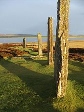 Cuatro grandes piedras verticales se sientan en un campo de hierba y brezo. Ellos son iluminados por la luz del sol rojizo y se proyectan largas sombras a la izquierda. Un lago y colinas bajas se encuentran más allá.