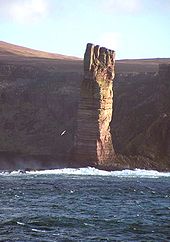 Una pila de altura perpendicular de roca de color marrón se encuentra en la luz del sol frente a una costa con acantilados que se encuentran en las sombras.