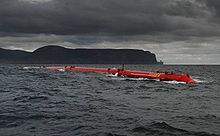 Un tubo largo de color rojo se encuentra en las aguas debajo, cielos cubiertos de nubes oscuras con negras colinas en la distancia.