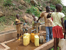 Fotografía que representa un adulto y cinco niños llenando bidones en una bomba de agua de metal rural con base de hormigón, en la parte inferior de una ladera rocosa escarpada