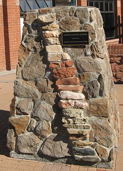 Un pilar robusto de la piedra de forma irregular motored con inserciones de apilado más roca en forma de ladrillo que forman una columna inclinada hacia la derecha. Una placa en el pilar lee: