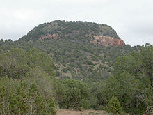 Un gran montículo de roca y tierra con color rojizo y el suelo gris y en su mayoría cubierto de vegetación.