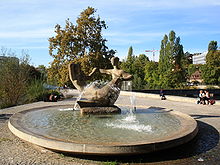 Fuente circular rodeado de pavimento. El centro de la fuente es una escultura de un par de figuras humanas abstractas.