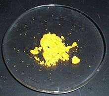 Algunos polvo amarillo canario se sienta, la mayoría en bultos, sobre un vidrio de reloj de laboratorio.