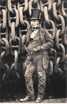 Un hombre del siglo 19 que lleva una chaqueta pantalón y chaleco, las manos en los bolsillos, el cigarro en la boca, que lleva un alto sombrero de copa de copa, de pie delante de cadenas de hierro gigantes en un tambor.
