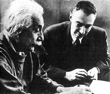 Einstein escrito en un escritorio. Oppenheimer se sienta a su lado, mirando.