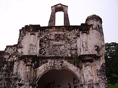 Ruina manchada de un edificio de piedra, mostrando un arco central, flanqueado por dos columnas, con un relieve de piedra por encima del arco, también flanqueado por dos columnas, y un segundo arco de sujeción independiente encaramado en lo más alto de la ruina.