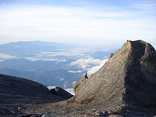 Una visión desde el pico de baja, con un pico más pequeño a la derecha de la foto, con montañas boscosas en el fondo