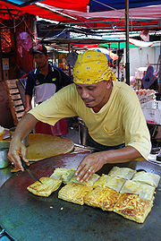 Un cocinero haciendo Murtabak, un tipo de panqueque, en un puesto al aire libre. Lo representan inclinando sobre su wok aplanada medida lleno de piezas de murtabak.