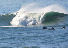 Foto de-alto-que-humano de tamaño ola rompiendo con varios surfistas viendo en primer plano