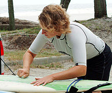 Foto de un hombre se inclinó sobre la tabla de surf barra de cera sólida roce contra el tablero con las palmeras y el océano en el fondo