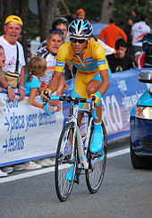 Un hombre vestido de amarillo y zapatos azules, andar en bicicleta, seguido por un coche. La gente lo está viendo desde detrás de una valla.