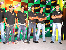 Un grupo de hombres de pie, vestidos con camisetas negras y azul vaqueros desgastados. Todos ellos están mirando a la izquierda de la imagen. The backdrop has alternate black and green boxes.