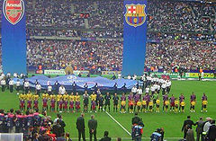 Dos equipos de deportistas, uno en camisas amarillas y una en camisas rojas y azules, de pie en una línea. En frente de la línea es una línea de niños con camisas equivalentes, pero cada uno en frente del equipo contrario. Un número de otras personas de pie en el primer plano mirando. Detrás de los equipos, un número de personas que tienen una gran pieza circular de tela con logotipos impresos, y dos grandes pancartas cuelgan verticalmente desde arriba toda la escena.