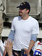 Hombre joven sonriente con una perilla de ajuste y el bigote, vestido con una camiseta blanca y una gorra de béisbol. Él está rodeado de manos llegar a él.