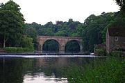 Un pont en pierre ?? deux arches ?? travers une rivi??re, vue le long de la rivi??re, les deux extr??mit??s cach??s par les arbres. Un d??versoir se trouve en face du pont, ?? l'extr??mit?? droite duquel se trouve un immeuble ?? deux ??tages.