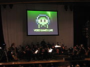 Une photographie d'un orchestre sur une sc??ne faiblement ??clair??e. Au-dessus du groupe est un ??cran de projection avec une image en noir, blanc et vert du pixel art. Le pixel art est un objet ovale portant un casque avec des yeux et quatre tentacules. Ci-dessous le pixel art est l'expression