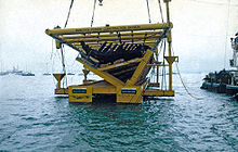 A partial section of a wooden ship lying inside a massive steel frame is suspended just about the water with a partial view of a barge on the left and small boats in the background.