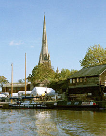 Un clocher d'??glise sur un quai avec des hangars et des bateaux couverts de b??ches en bois. En face de ces derniers sur l'eau un jumeau mat bateau ?? voile et un narrowboat