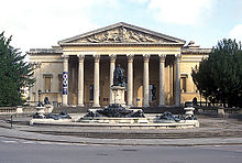 A Palladian style nineteenth century stone building with a large colonnaded porch. In front a large metal statue on a pedestal and fountains with decorations.