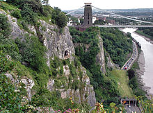 Rocky c??t?? d'une gorge avec une plate-forme en face d'une grotte ?? mi-hauteur. A droite sont une route et la rivi??re. Dans la distance sont un pont et b??timents suspension.