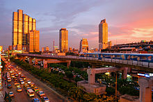 A road with a lot of traffic; an elevated train running above the road; several buildings, one with the appearance of a robot, during sunset