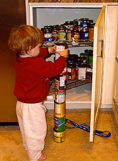 Jeune gar??on roux dos ?? la cam??ra, l'empilage septi??me bo??te sommet d'une colonne de six bo??tes de conserve sur le plancher de la cuisine. Un cellier ouverte contient beaucoup plus de canettes.